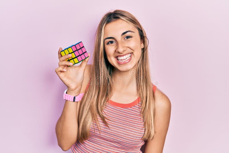 Beautiful hispanic woman holding colorful puzzle cube intelligence game looking positive and happy standing and smiling with a stock photo