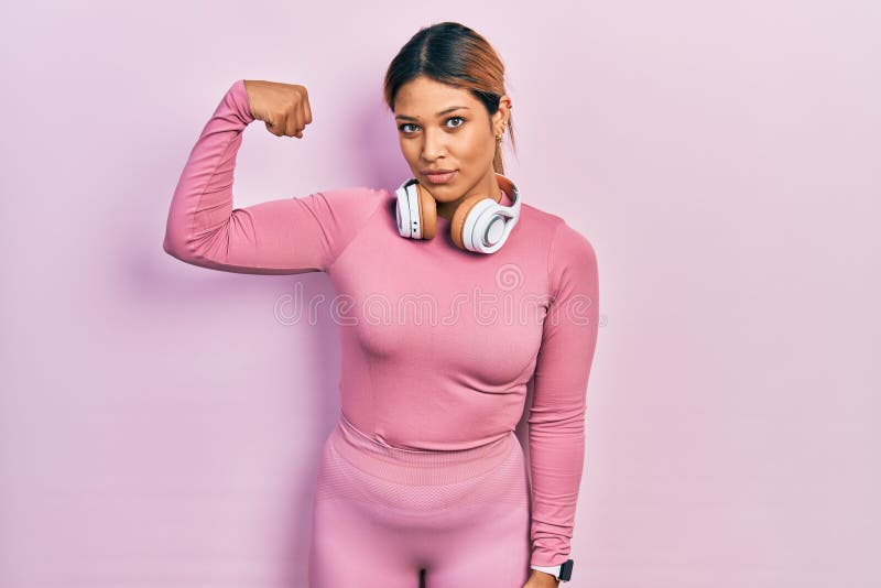 Beautiful hispanic woman wearing gym clothes and using headphones strong person showing arm muscle, confident and proud of power