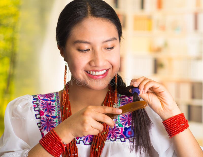 Beautiful hispanic woman wearing white blouse with colorful embroidery, using small hairbrush during makeup routine, garden background.