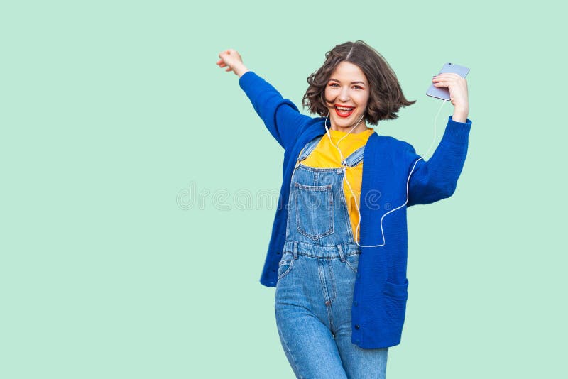 Beautiful joyful young adult woman in hipster wear in denim overalls standing, holding phone and listening favourite music with