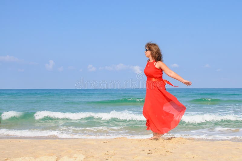 Beautiful lady in bright elegant red dress at beach, with sea and blue sky as background