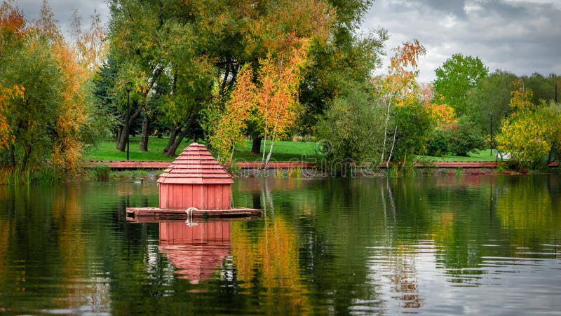 Beautiful lake with small wooden houses for ducks and autumn willow trees with green, yellow leaves in city park. Moscow