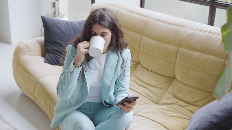 Beautiful Latina businesswoman drinks coffee during a break, sitting on a sofa while using a smartphone