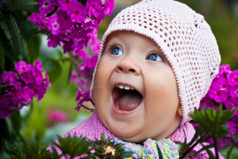 A beautiful little girl with big blue eyes in pink knitted hat is happy and laughing among the red flowers in the garden royalty free stock photography A beautiful little girl with big blue eyes in pink knitted hat is happy and laughing among the red flowers in the garden royalty free stock photography
