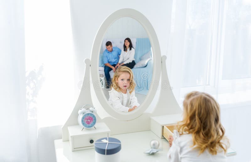 Beautiful little girl with blonde long hair, looking at herself in mirror indoors. Mother and father is in reflection royalty free stock photo Beautiful little girl with blonde long hair, looking at herself in mirror indoors. Mother and father is in reflection royalty free stock photo