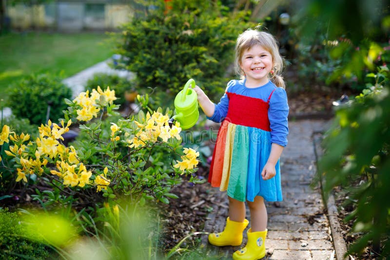 Beautiful little toddler girl in yellow rubber boots and colorful dress watering spring flowers with kids water can