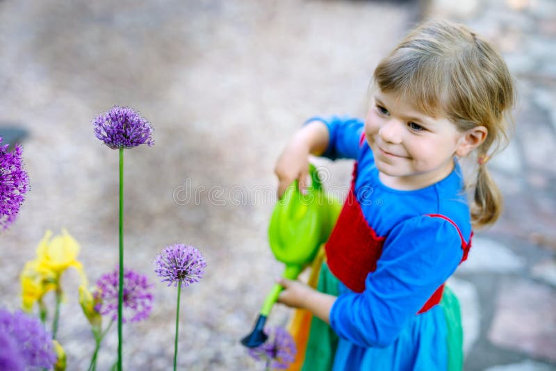 Beautiful little toddler girl in yellow rubber boots and colorful dress watering spring flowers with kids water can