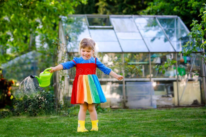 Beautiful little toddler girl in yellow rubber boots and colorful dress watering spring flowers with kids water can