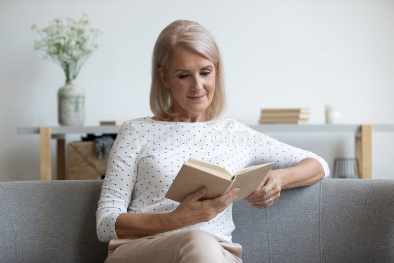 Beautiful mature woman reading book, sitting on couch at home