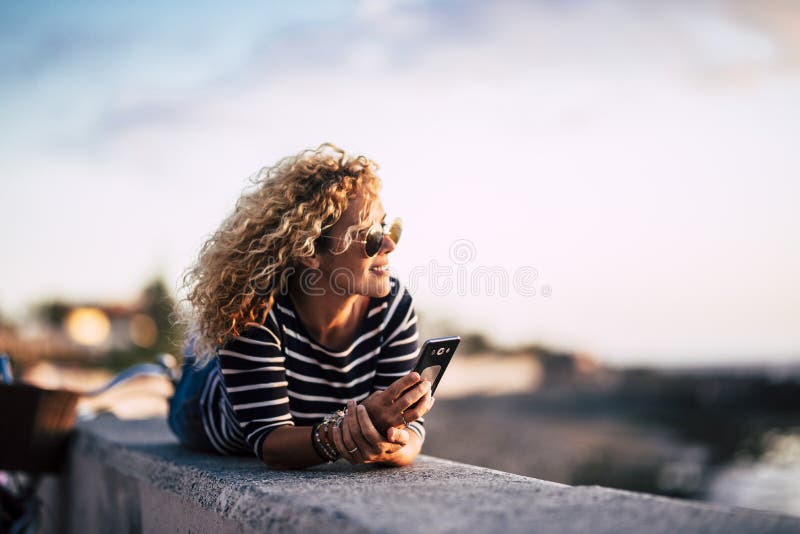 Beautiful middle age blonde curly womoan enjoy and relax in outdoor leisure activity looking at the sunset with modern phone ready stock photos Beautiful middle age blonde curly womoan enjoy and relax in outdoor leisure activity looking at the sunset with modern phone ready stock photos