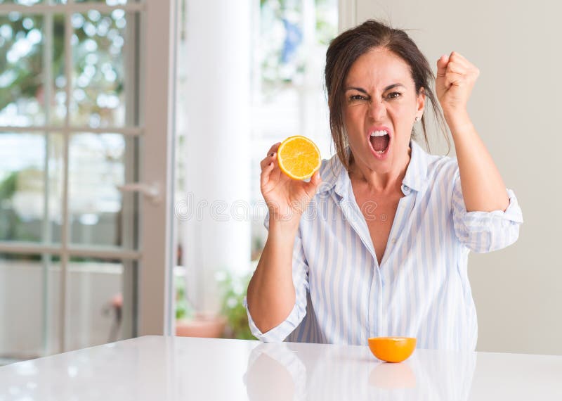 Middle aged woman holding orange fruit annoyed and frustrated shouting with anger, crazy and yelling with raised hand, anger concept. Middle aged woman holding orange fruit annoyed and frustrated shouting with anger, crazy and yelling with raised hand, anger concept