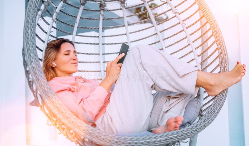 Beautiful middle-aged woman listening Earbuds smartphone music lying in comfortable hanging chair on open house terrace and