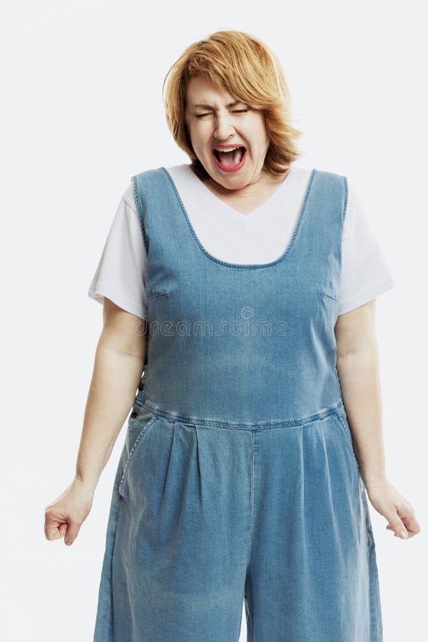 A beautiful middle-aged woman with red hair screams emotionally. Dressed in denim overalls. White background. Vertical.