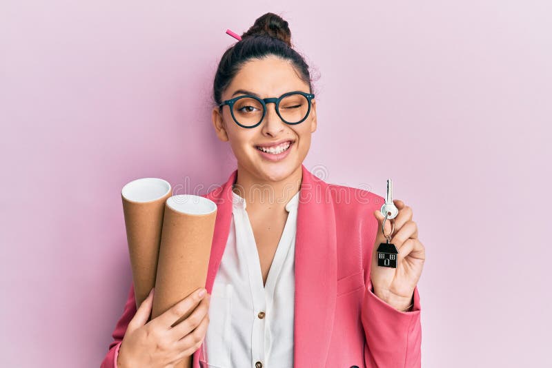 Beautiful middle eastern woman holding paper blueprints and house keys winking looking at the camera with sexy expression