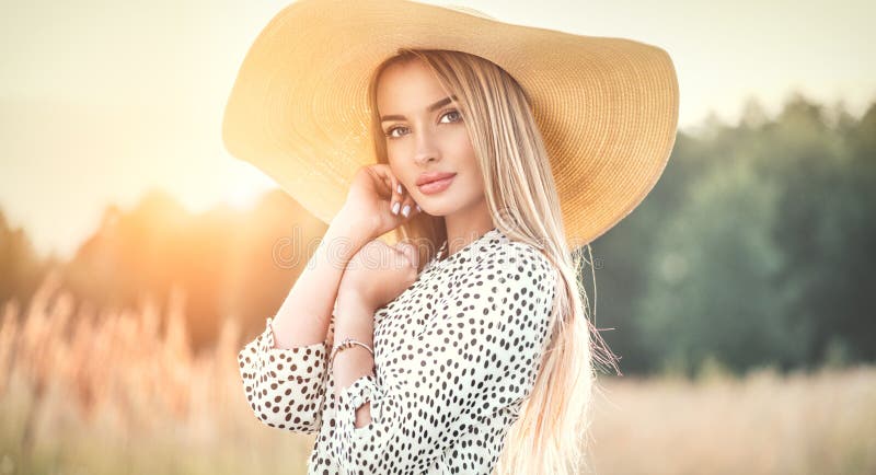 Beautiful model girl posing on a field, enjoying nature outdoors in wide brimmed straw hat. Beauty blonde young woman