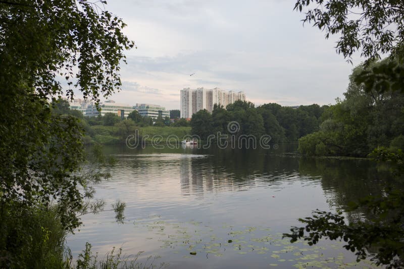 Beautiful nature with pond lake, green trees foliage, grass, buildings and clouds in the background. Afternoon panorama landscape