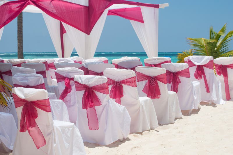 Beautiful outdoor wedding on beach scene: white chairs and arch decorated with red ribbon