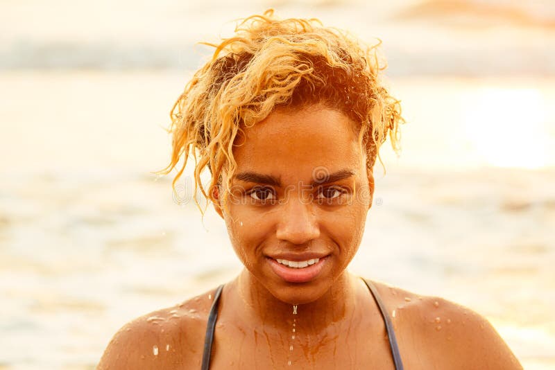 Beautiful portrait of african american surfer woman with wet afro blonde curly hair in bikini at sunset on beach.