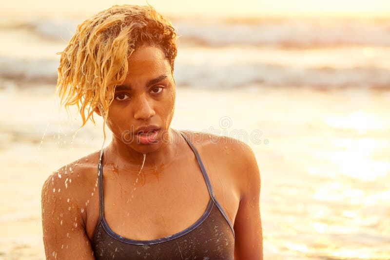 Beautiful portrait of african american surfer woman with wet afro blonde curly hair in bikini at sunset on beach.