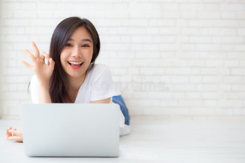 Beautiful portrait asian young woman working online laptop with excited lying on floor brick cement background, freelance girl