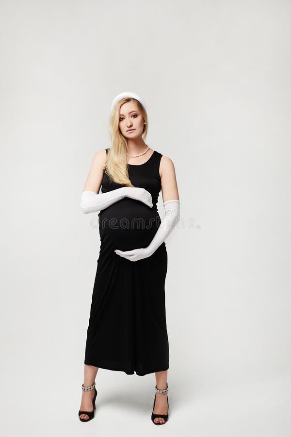 Beautiful pregnant woman in black evening dress and long white gloves touching her belly and posing on a white