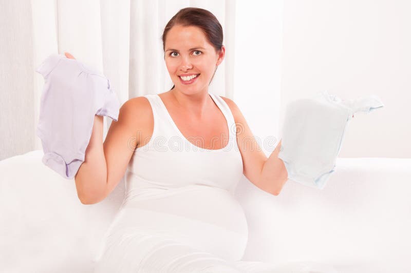 Beautiful pregnant woman holding baby clothes- bodysuits