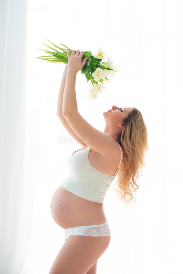 Beautiful pregnant woman with spring white flowers in front of a window flooded with sunlight. Female health and happy pregnancy stock photography Beautiful pregnant woman with spring white flowers in front of a window flooded with sunlight. Female health and happy pregnancy stock photography
