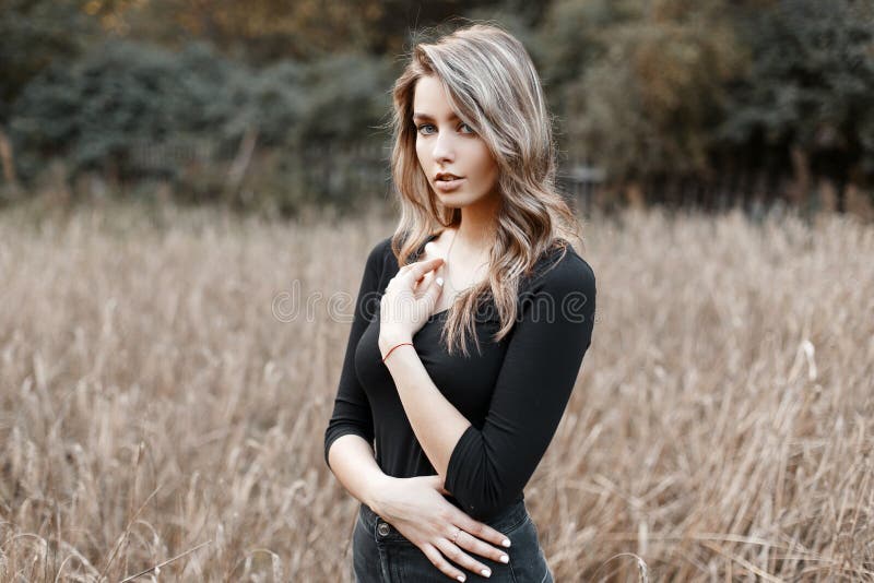 Beautiful pretty sexy young woman in a black stylish T-shirt with blond hair in trendy jeans poses in a field on a warm autumn day