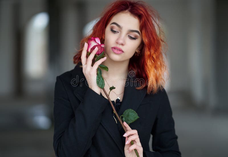 Beautiful red-haired girl in a black coat holding a pink rose in runes and touching the flower to the face