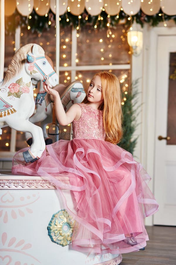 Beautiful red-haired girl with long hair and blue eyes rides a carousel in a long pink dress. Carousel in the form of horses