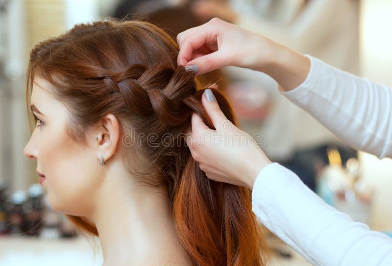 Beautiful, red-haired girl with long hair, hairdresser weaves a French braid, in a beauty salon