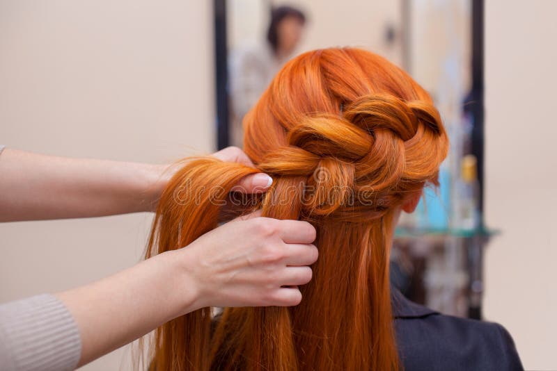 Beautiful, red-haired girl with long hair, hairdresser weaves a French braid, in a beauty salon