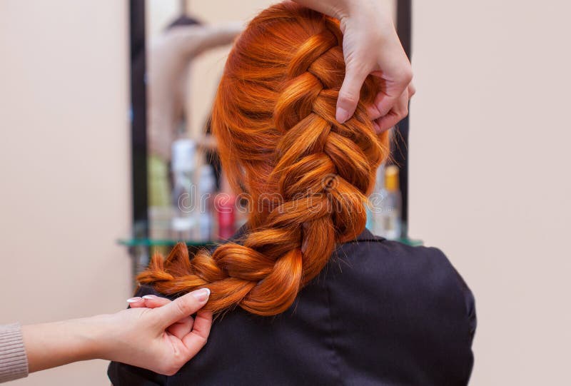 Beautiful, red-haired girl with long hair, hairdresser weaves a French braid, in a beauty salon