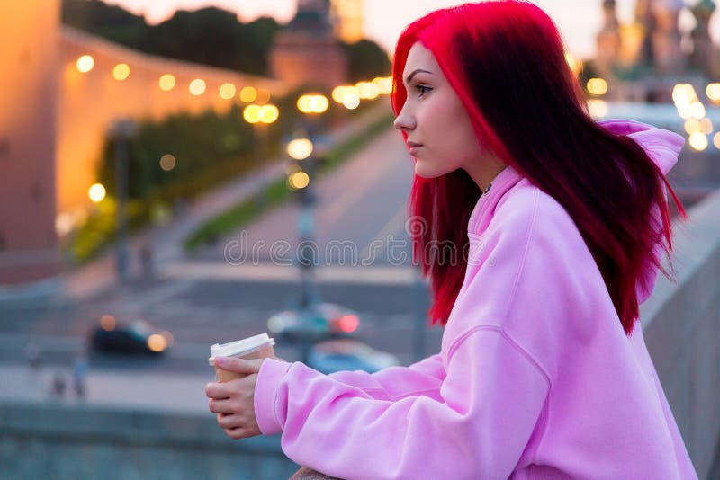 Krásný dospívající v růžový mikina pití káva v na osvětlené město ulice.