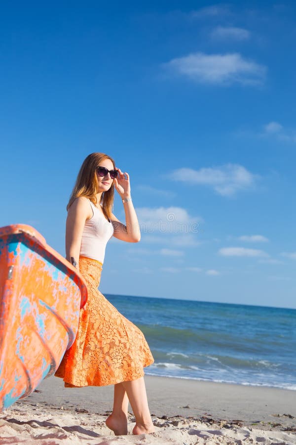Beautiful red-haired young woman in sunglasses and skirt relaxing on beach near orange boat. stock photo Beautiful red-haired young woman in sunglasses and skirt relaxing on beach near orange boat. stock photo
