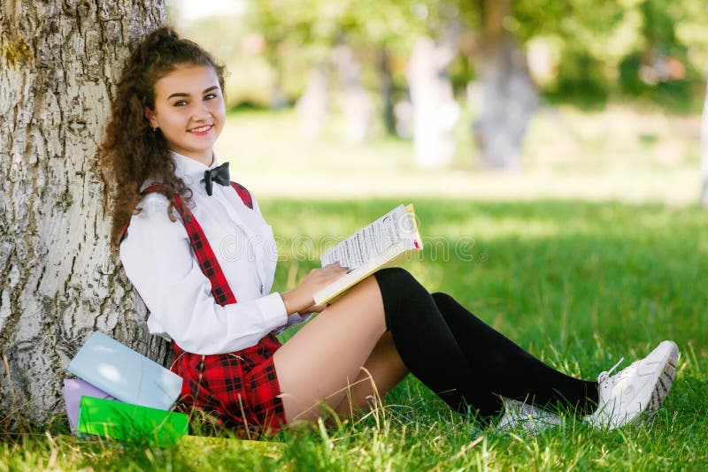 Beautiful European Schoolgirl Sits In A Park Near A Tree On The Green 