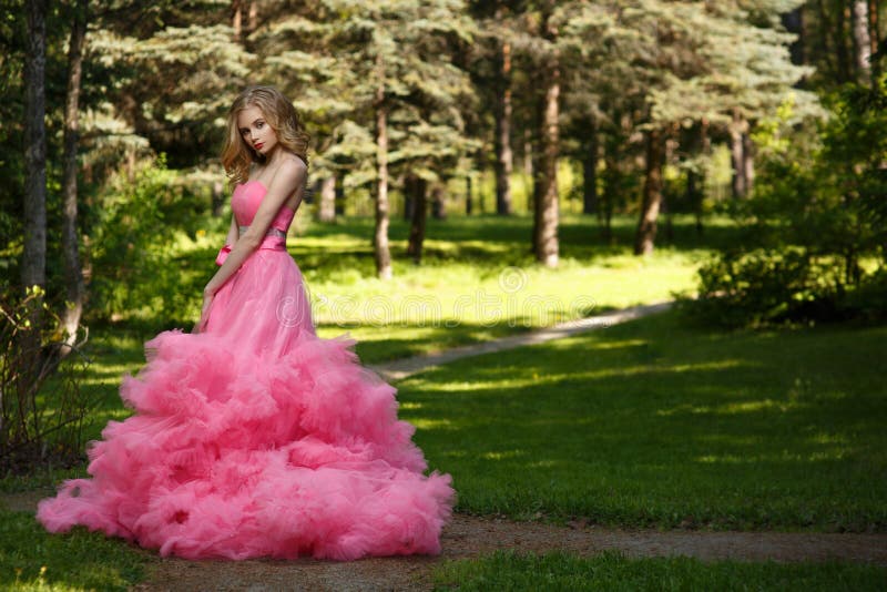 Sensual woman in pink evening dress with fluffy skirt is posing in botanical garden on the grass surrounded by the woods