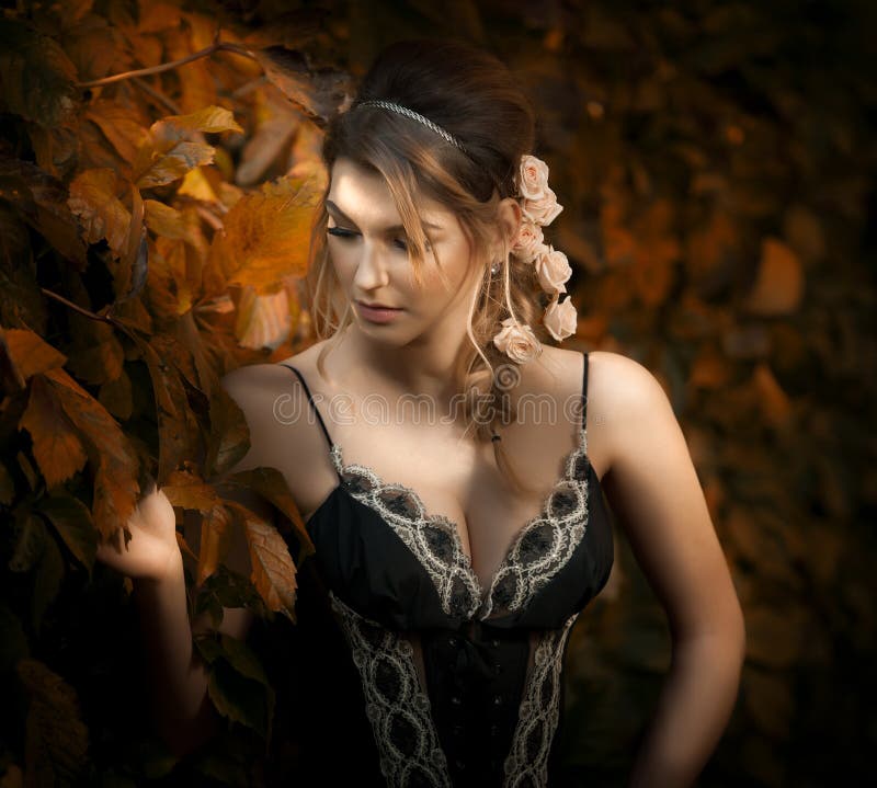 Beautiful sensual woman with roses in hair posing near a wall of green leaves. Young female in black elegant dress daydreaming