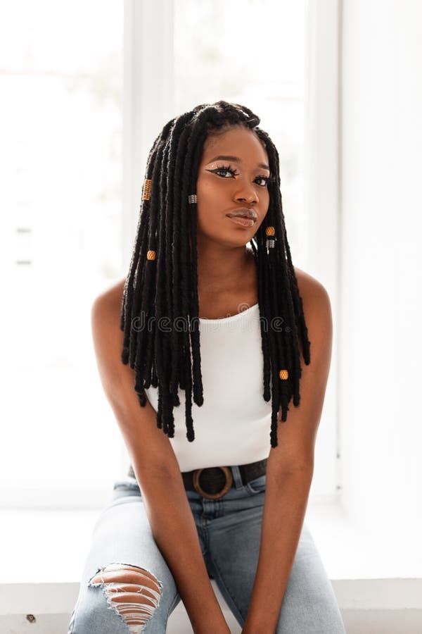 Beautiful sexy American black woman with stylish dreadlocks in fashion denim clothes near window in room on bright day. Fashion