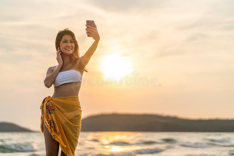 Beautiful sexy Asian girl smiling taking selfie photo or live video call, using smartphone on beach vacation at sunset royalty free stock image Beautiful sexy Asian girl smiling taking selfie photo or live video call, using smartphone on beach vacation at sunset royalty free stock image
