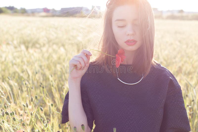 Beautiful cute girl with big lips and red lipstick in a black jacket with a flower poppy standing in a poppy field at sunset