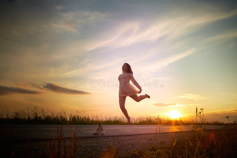 Beautiful sexy girl in pink dress jumping on the asphalt highway during sunset and field with grass on the side of the road