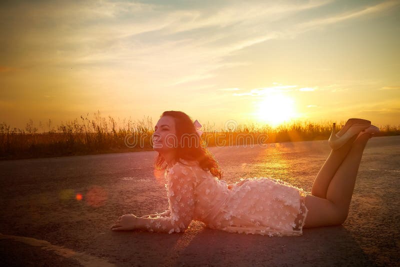 Beautiful sexy girl in pink dress lying on the asphalt highway during sunset and field with grass on the side of the road