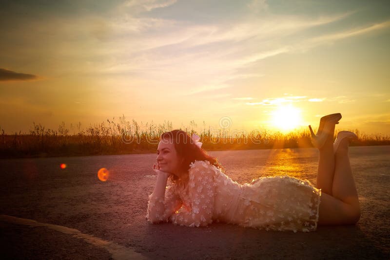 Beautiful sexy girl in pink dress lying on the asphalt highway during sunset and field with grass on the side of the road