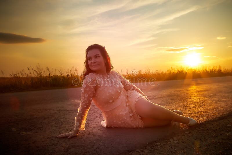 Beautiful sexy girl in pink dress sitting on the asphalt highway during sunset and field with grass on the side of the road