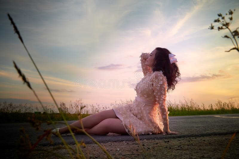 Beautiful sexy girl in pink dress sitting on the asphalt highway during sunset and field with grass on the side of the road