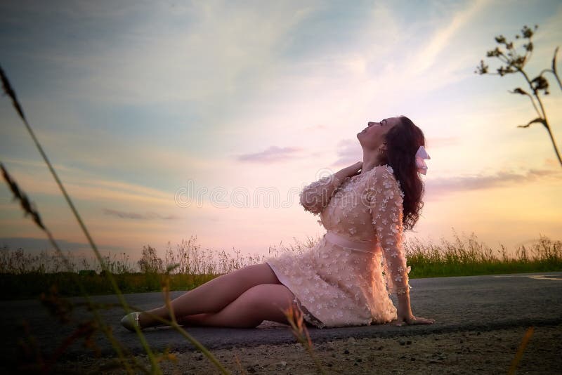 Beautiful sexy girl in pink dress sitting on the asphalt highway during sunset and field with grass on the side of the road