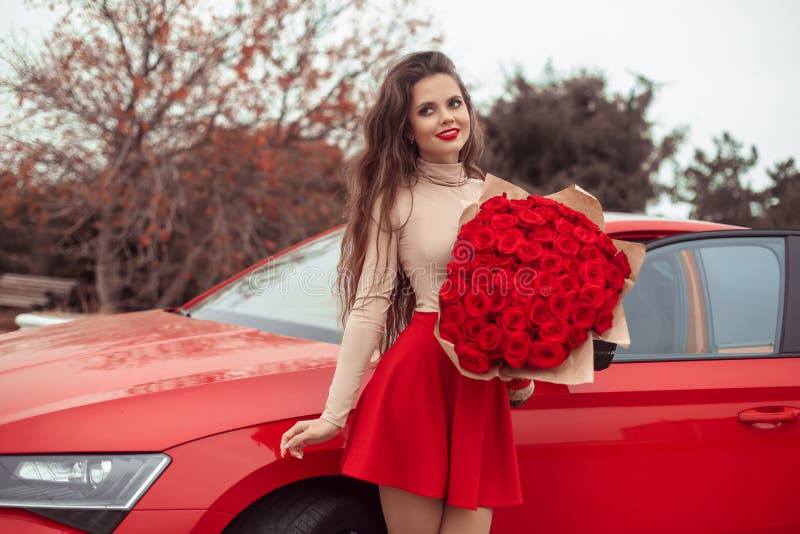 Beautiful sexy woman with rose bouquet of flowers posing by new red car. Charming brunette in red skirt. Beauty outdoor autumn