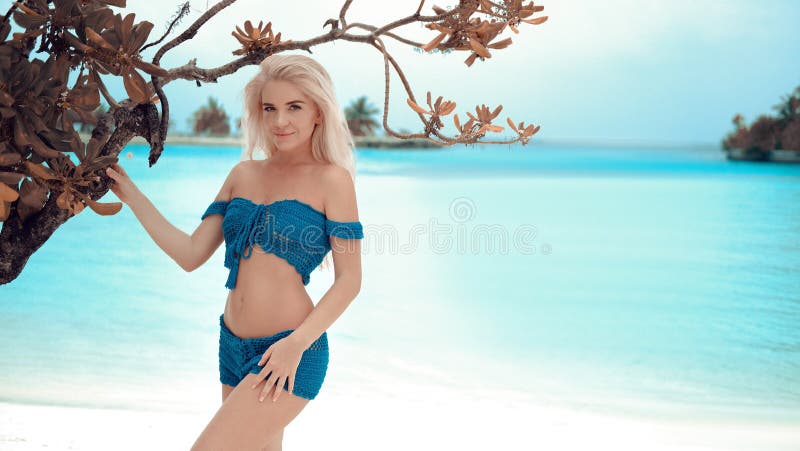 Beautiful sexy woman wearing white Crochet bikini swimwear posing by tree on turquoise ocean lagoon.  Slim blonde model resting on Maldives. Summer vacations