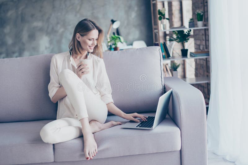 Beautiful smiling blonde woman sitting on couch using wi fi internet on her laptop for online shopping holding cup of tea